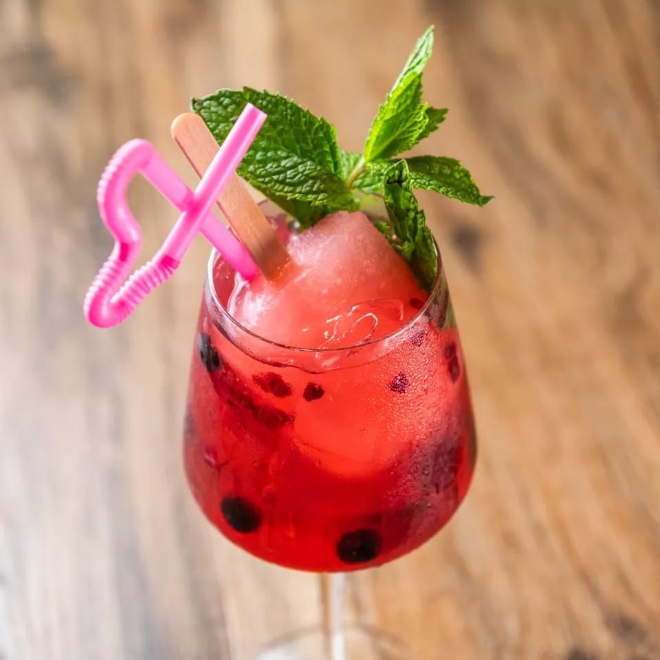 Red cocktail with blueberries, mint, a pink straw, and a popsicle on a wooden table.
