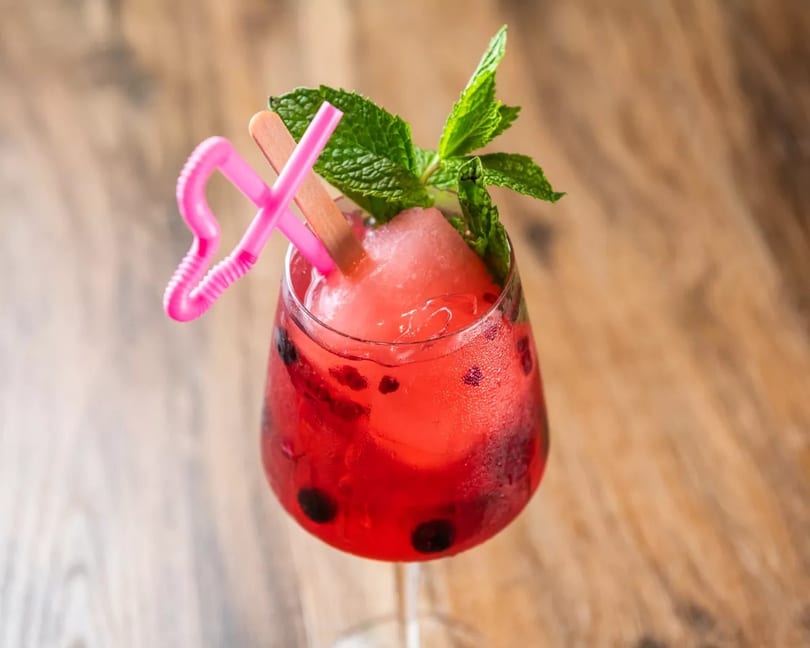 Red cocktail with blueberries, mint, a pink straw, and a popsicle on a wooden table.