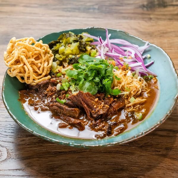 A vibrant bowl of Khao Soi with braised beef, crispy noodles, cilantro, and red onions.