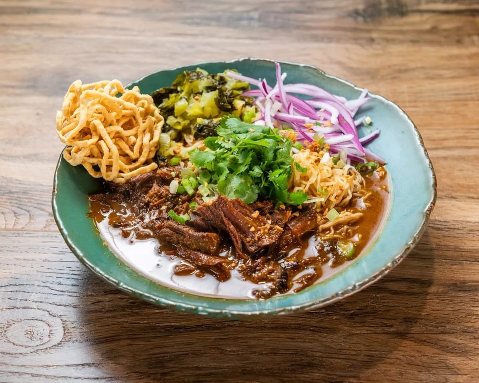 A vibrant bowl of Khao Soi with braised beef, crispy noodles, cilantro, and red onions.