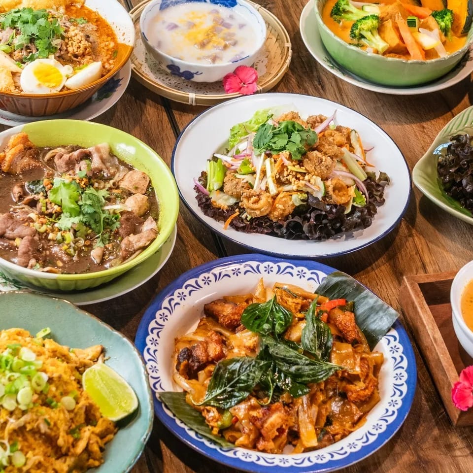 A wooden table laden with various colorful Thai dishes, including noodles, curries, and salads.