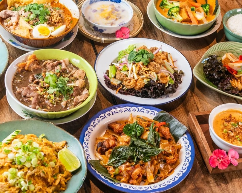 A wooden table laden with various colorful Thai dishes, including noodles, curries, and salads.