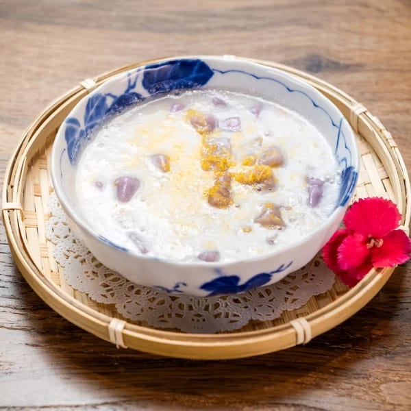 A bowl of taro dessert soup with purple taro chunks and yellow topping, on a bamboo tray.