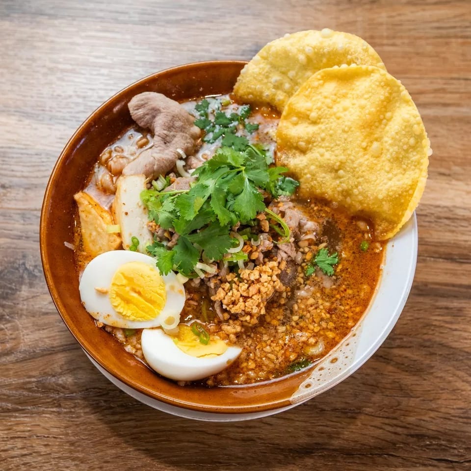 Spicy Thai noodle soup with pork, egg, cilantro, and crispy wontons in a brown bowl.