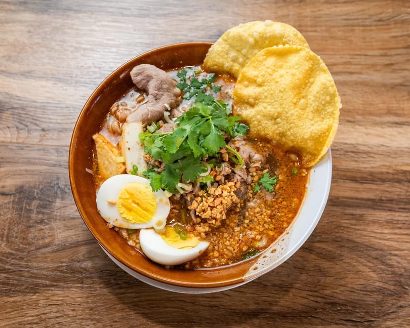 Spicy Thai noodle soup with pork, egg, cilantro, and crispy wontons in a brown bowl.