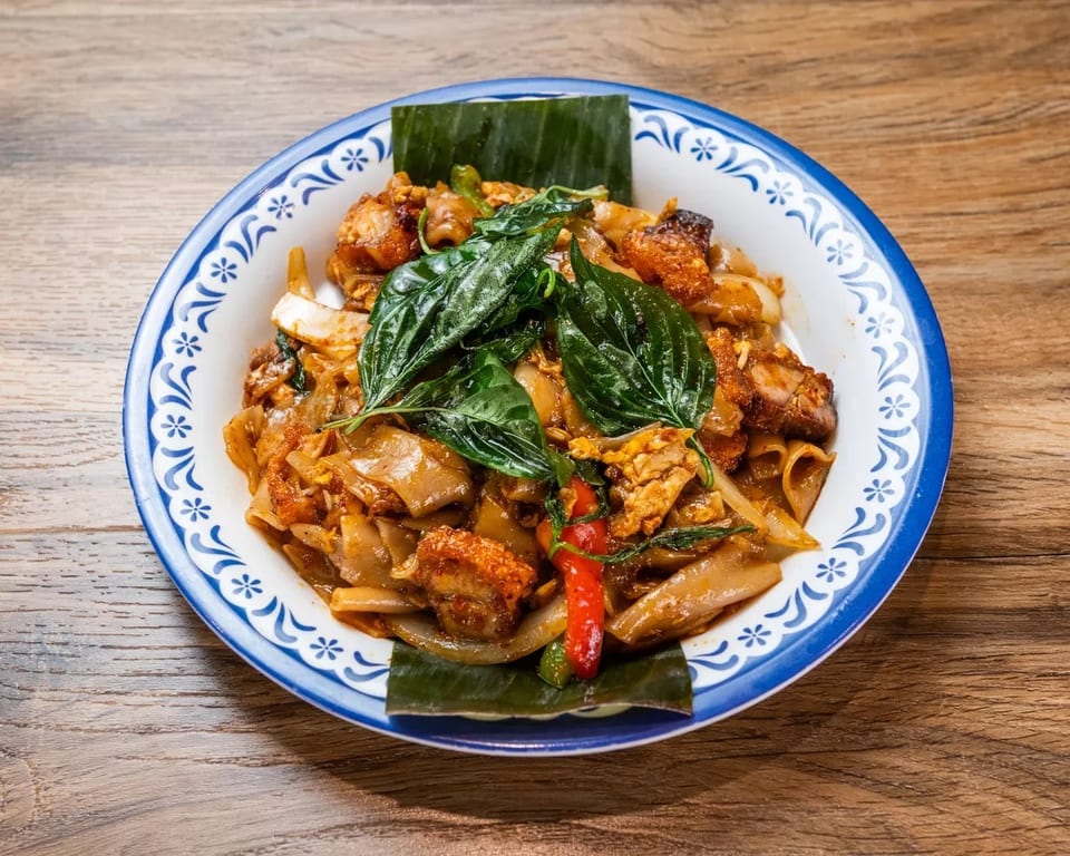 Pad See Ew with crispy pork belly, wide noodles, basil, and chili on a decorative plate.