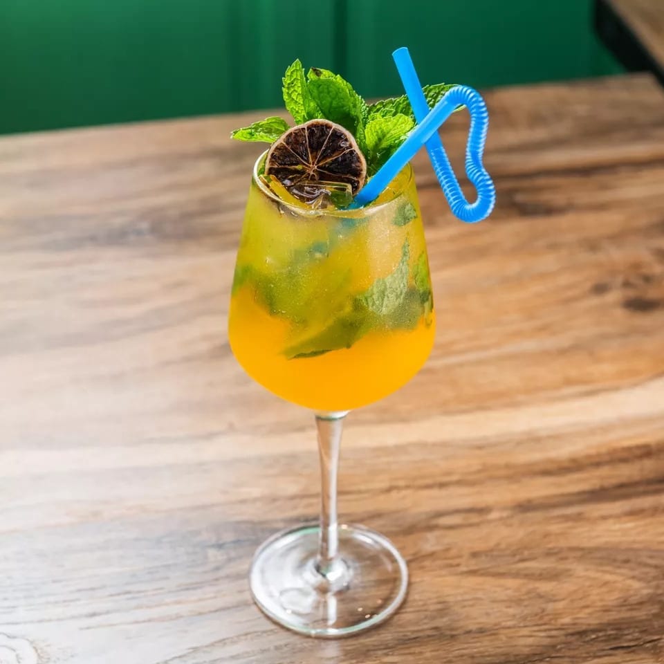 A vibrant yellow and green cocktail with mint, a dried orange slice, and a blue straw.