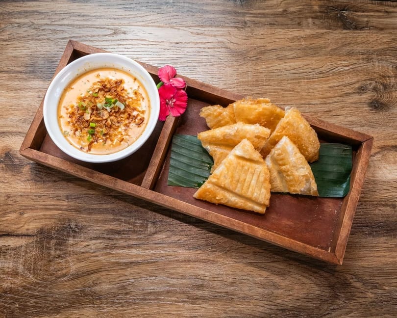 Fried triangular pastries and a bowl of dipping sauce with fried onions and scallions on a wooden tray.