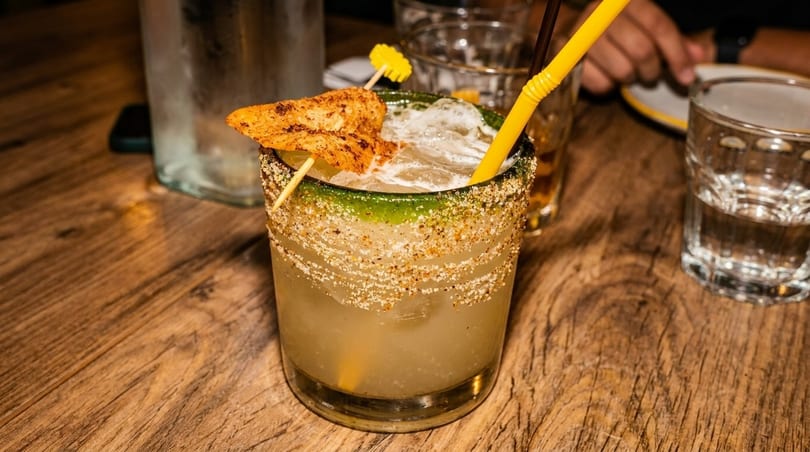 A margarita with a green salted rim, yellow straw, and a chili-dusted mango garnish on a wooden table.